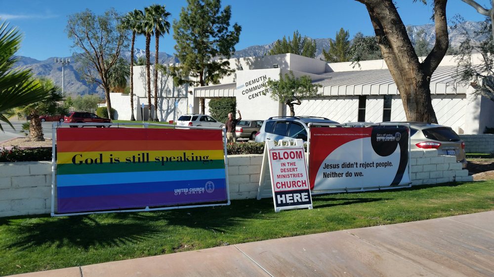 Rainbow flag placed by the sidewalk in front of a church that says "God is still speaking"