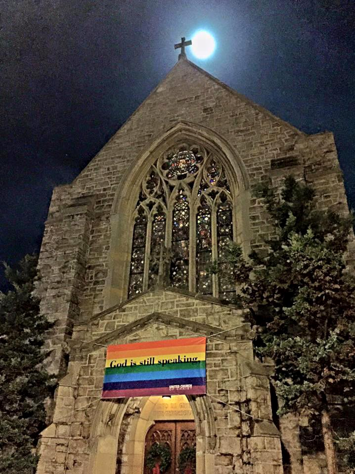 Rainbow sign above the entrance of a church that says "God is still speaking"